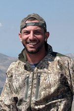 Ethan Proud smiling in front of mountains wearing camouflage jacket and backwards baseball cap.