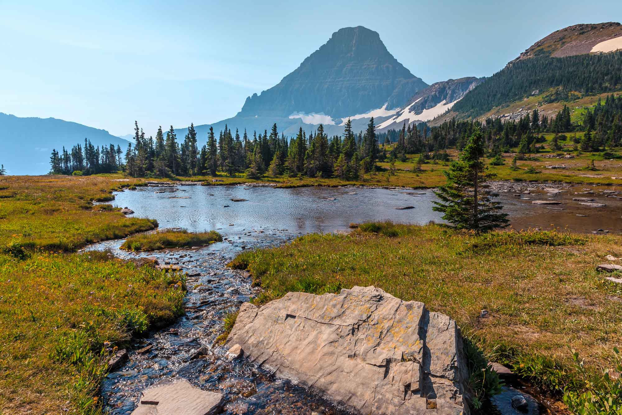 Crown of the Continent ecosystem: stream flows into pond with coniferous trees in midground and iconic mountain peak in background