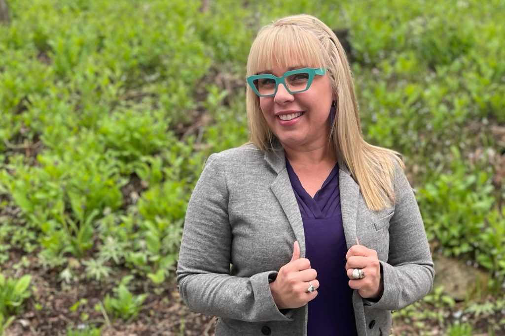 a woman stands in front of emerging spring plants; the woman is white with long, blonde hair; she wears a gray blazer over a purple shirt and thick-rimmed teal glasses